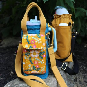 May include: Two water bottle carriers. The first is teal with a mustard yellow strap and a mustard yellow pocket with a floral design. The second is mustard yellow with a black strap. Both have a handle and a shoulder strap.