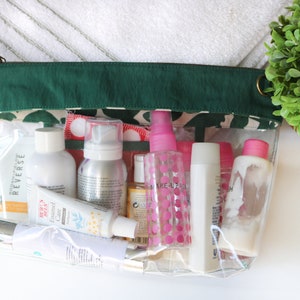 May include: A clear plastic zippered pouch with a green fabric top, filled with various travel-sized toiletries, including bottles, tubes, and sprays. The pouch is sitting on a white surface.