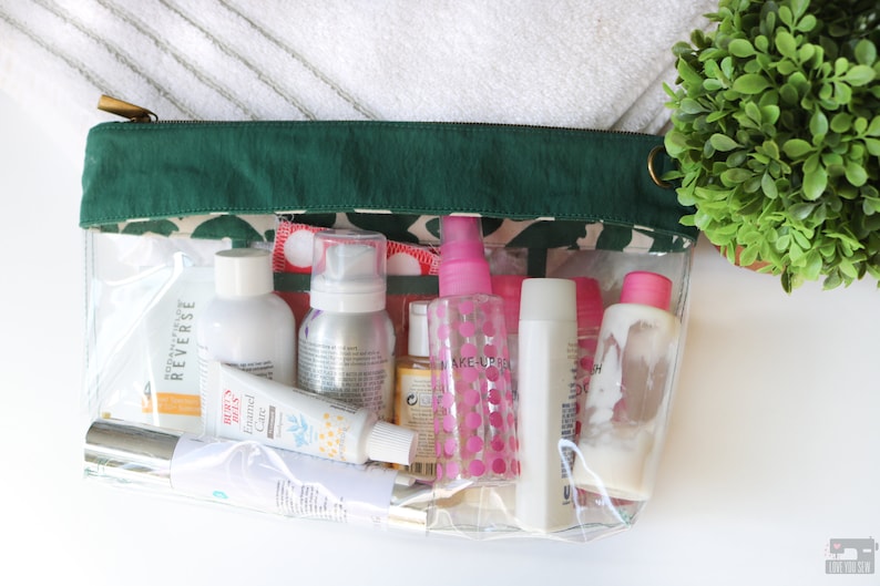 May include: A clear plastic zippered pouch with a green fabric top, filled with various travel-sized toiletries, including bottles, tubes, and sprays. The pouch is sitting on a white surface.