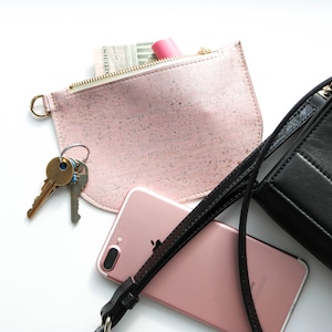 Puede incluir: Una cartera de corcho rosa con cierre de cremallera, una correa de cuero negro, un llavero dorado con llaves, un pintalabios rosa y un iPhone rosa dorado.