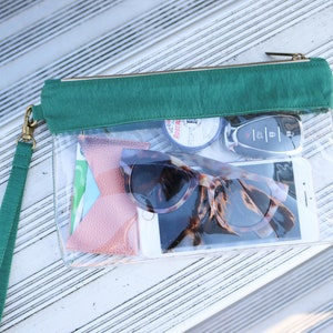 May include: A clear plastic zippered pouch with a green fabric flap. The pouch contains a pair of sunglasses, a cell phone, a key fob, and other small items.