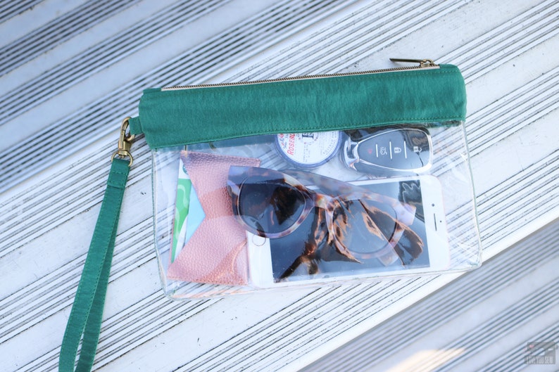 May include: A clear plastic zippered pouch with a green fabric flap. The pouch contains a pair of sunglasses, a cell phone, a key fob, and other small items.