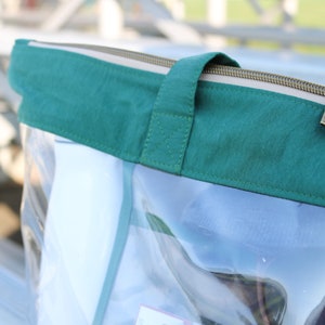 May include: Clear plastic tote bag with a green fabric top and a zipper closure. The zipper has a gold pull tab.