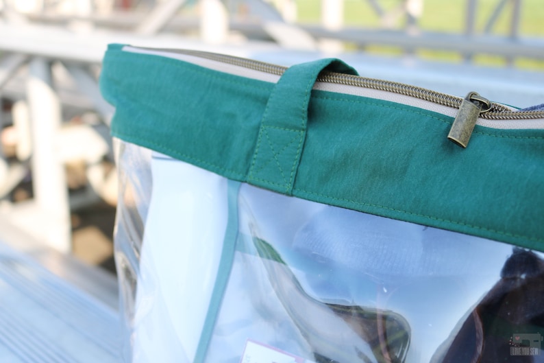 May include: Clear plastic tote bag with a green fabric top and a zipper closure. The zipper has a gold pull tab.