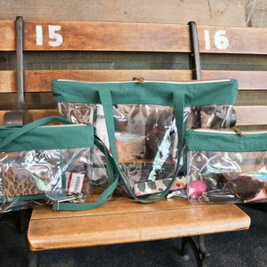 May include: Three clear plastic tote bags with green fabric trim and straps. The bags are lined up on a wooden bench. The largest bag is a tote bag, the middle bag is a crossbody bag, and the smallest bag is a clutch. The bags are filled with various items, including a phone, wallet, sunglasses, and a pink ball.