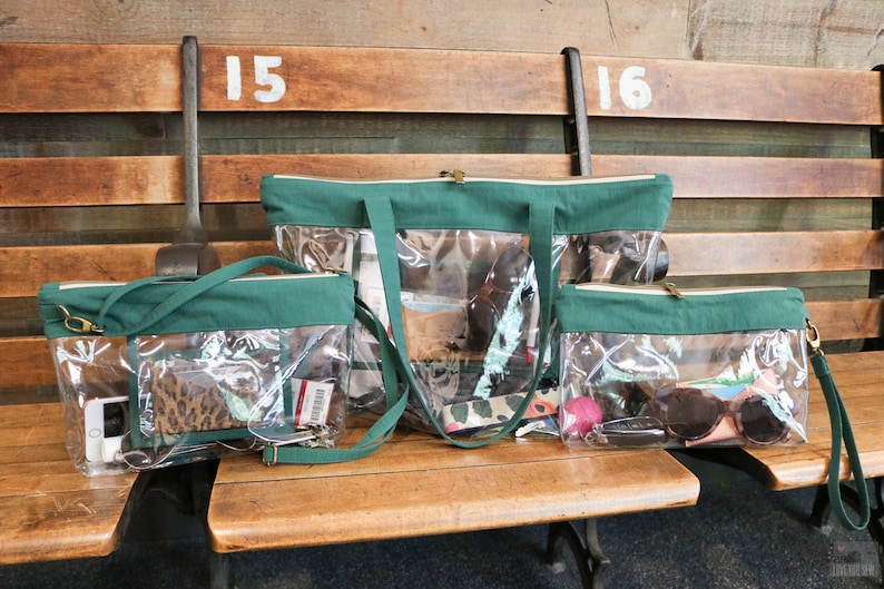 May include: Three clear plastic tote bags with green fabric trim and straps. The bags are lined up on a wooden bench. The largest bag is a tote bag, the middle bag is a crossbody bag, and the smallest bag is a clutch. The bags are filled with various items, including a phone, wallet, sunglasses, and a pink ball.