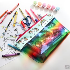May include: A clear rainbow-colored zippered pouch with a unicorn design and a rainbow and cloud pattern. The pouch is filled with crayons, markers, and colored pencils.