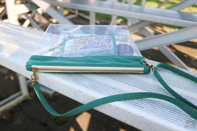 May include: A clear plastic zippered pouch with a teal green strap and a teal green fabric flap. The pouch is lying on a white metal bench and has a gold zipper pull.