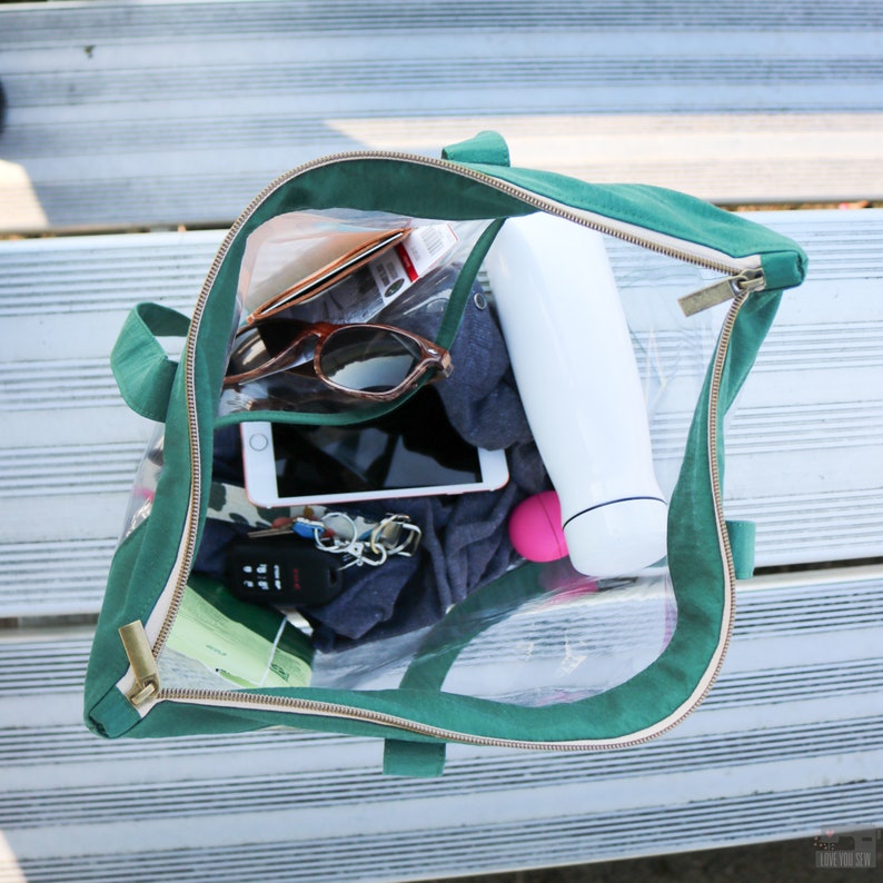May include: A clear green tote bag with a zipper closure. The bag is open and filled with various items, including a tablet, sunglasses, keys, a water bottle, and a small pink container.