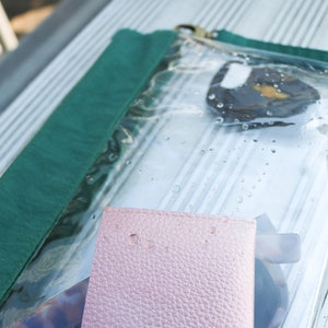 May include: A clear plastic zippered pouch with a green fabric flap. The pouch contains a pink leather wallet, a pair of sunglasses, and a small round object.