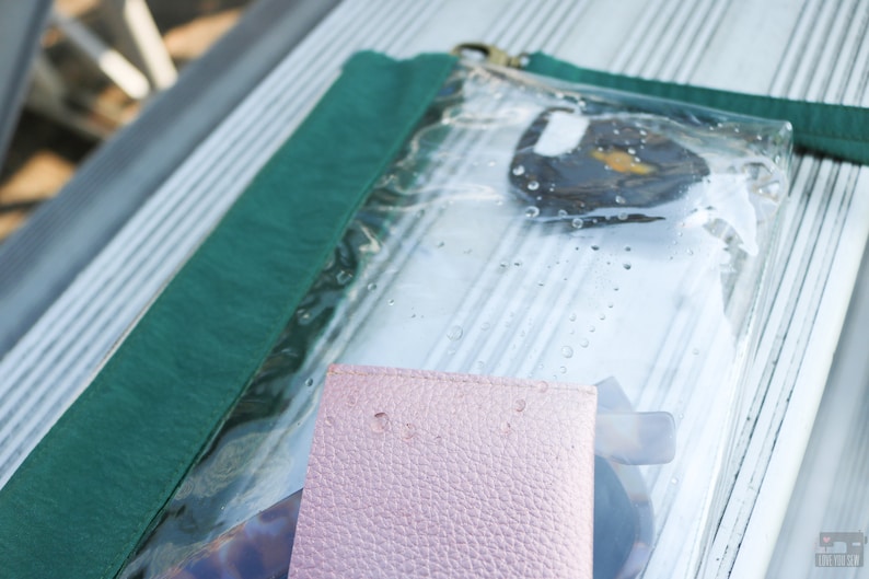 May include: A clear plastic zippered pouch with a green fabric flap. The pouch contains a pink leather wallet, a pair of sunglasses, and a small round object.
