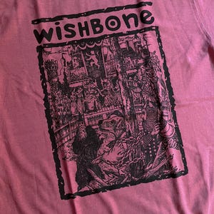 May include: A dusty rose-colored t-shirt with a black graphic. The graphic features the word "wishbone" above a detailed black and white illustration of a medieval scene with figures and a chained bear. The shirt has a crew neck.