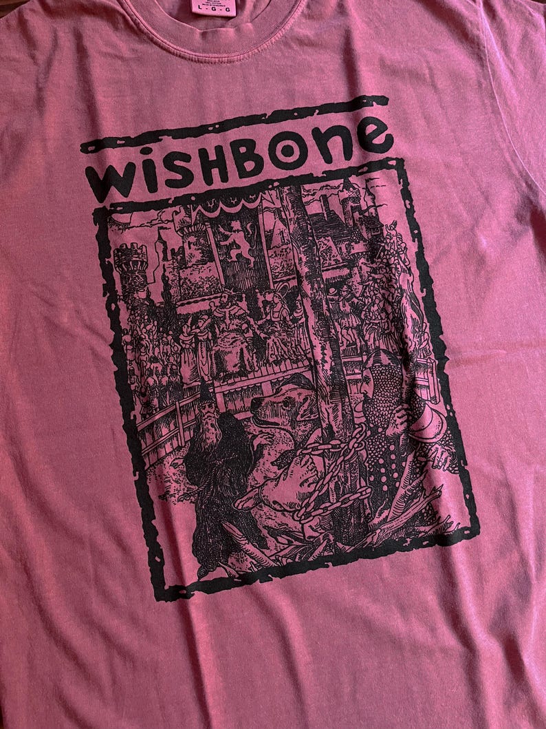 May include: A dusty rose-colored t-shirt with a black graphic. The graphic features the word "wishbone" above a detailed black and white illustration of a medieval scene with figures and a chained bear. The shirt has a crew neck.
