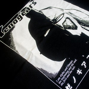 May include: Black and white graphic t-shirt with the text "Xenogears" and a silhouette of a character. The text below the character reads "I am Alpha and Omega, the beginning and the end, the first and the last. ゼノギアス"