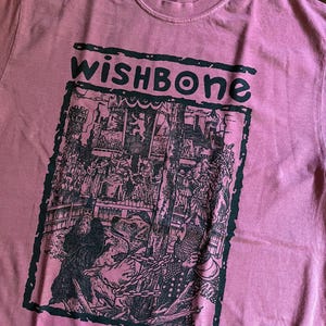 May include: A dusty rose-colored t-shirt with the word "wishbone" in dark green block letters. Below the text is a black and white graphic illustration of a medieval scene with figures and a chained bear.