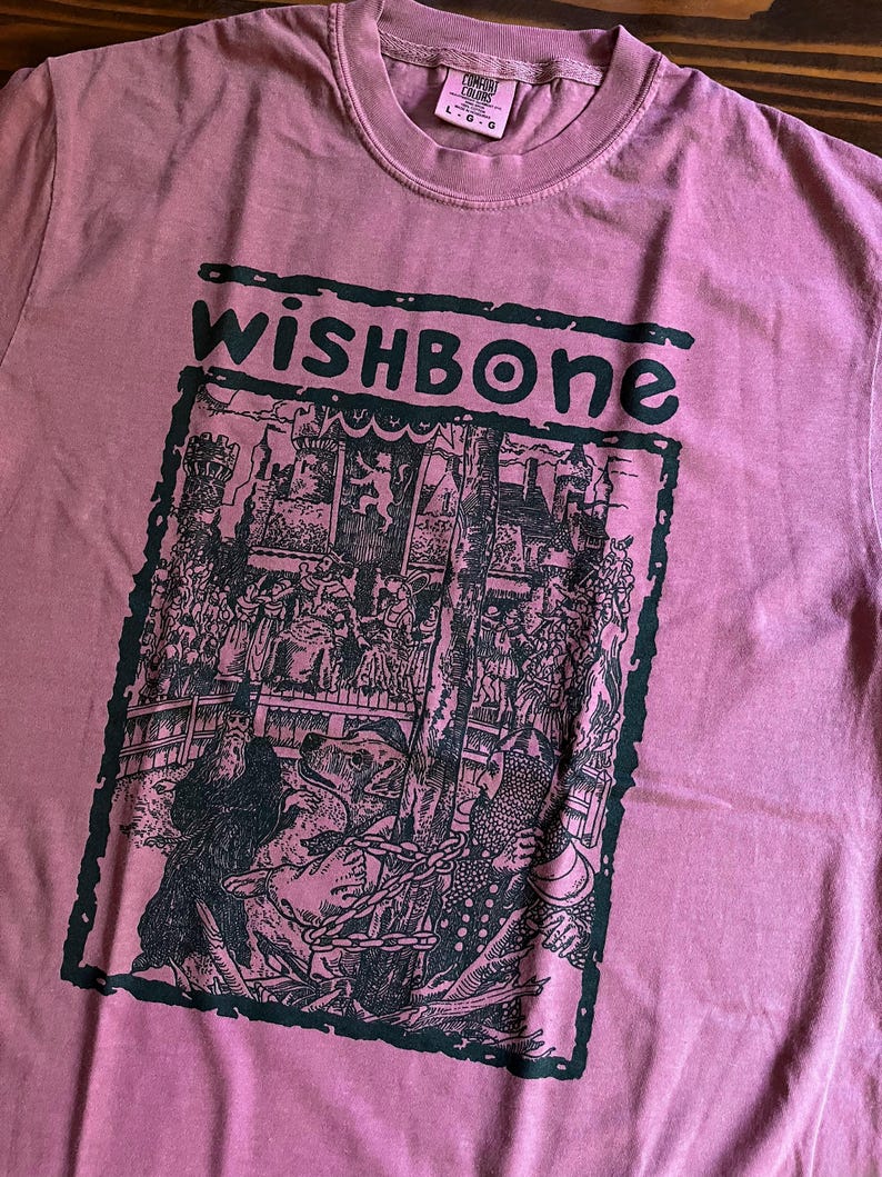 May include: A dusty rose-colored t-shirt with the word "wishbone" in dark green block letters. Below the text is a black and white graphic illustration of a medieval scene with figures and a chained bear.