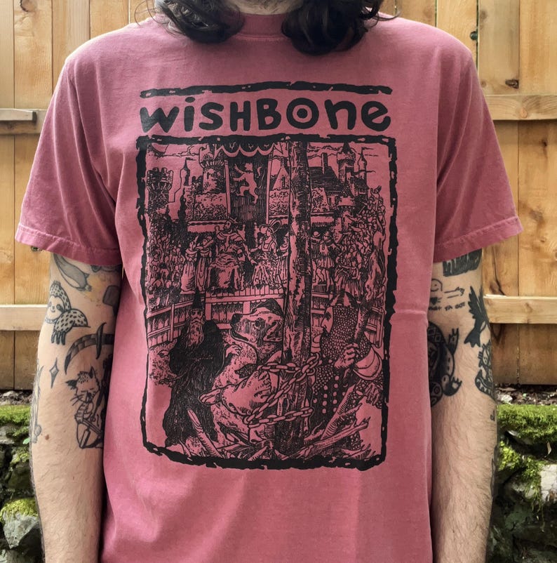 May include: A dusty rose-colored t-shirt with the word "WISHBONE" in a black, stylized font. The graphic depicts a detailed black and white illustration of a medieval scene with figures and architecture.