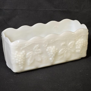 May include: A rectangular, off-white glass dish with a scalloped rim. The sides are decorated with embossed grape clusters and leaves. The dish has a rectangular shape and is likely used for serving or display.