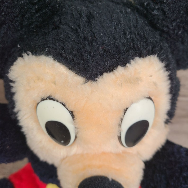 May include: Close-up of a plush Mickey Mouse toy with black fur, large white eyes, and a beige face.
