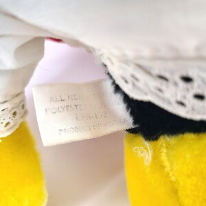 Pu&ograve; includere: Primo piano di un'etichetta bianca con testo nero che recita "All New Material Polyester", attaccata a un giocattolo di peluche giallo, nero e bianco.