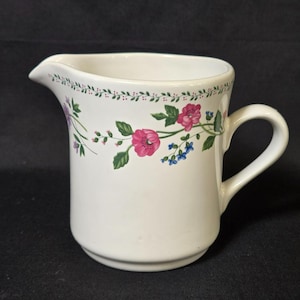 May include: White ceramic creamer with a floral design. The creamer features a spout and a handle. The floral pattern includes pink flowers, green leaves, and small blue flowers. A green and red dotted border decorates the rim.