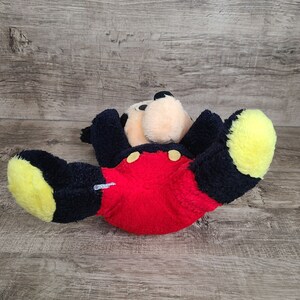 May include: A plush Mickey Mouse toy with black fur, red shorts with yellow buttons, and yellow ears. The toy is lying on its back with its arms and legs spread out.