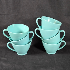 May include: A set of six solid light turquoise cups with a smooth finish, each featuring a single curved handle and a flared rim. The cups are arranged in two stacks, with three cups in each stack, showcasing a consistent color and design.