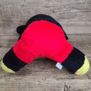May include: Black and red plush toy with yellow accents. The toy is shaped like a cartoon character and is lying on its stomach. The toy has a tag that reads "Made in China".