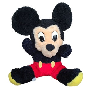 May include: A plush black and red Mickey Mouse toy with large black ears and a big smile. The toy has yellow accents on its feet.