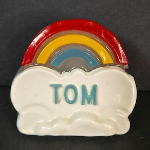 May include: A colorful rainbow-shaped toy with the name "TOM" in teal letters on a white cloud. The rainbow features red, yellow, and blue arches. The toy is made of plastic and has a glossy finish.
