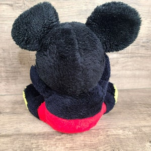 May include: A black plush Mickey Mouse toy with red shorts and yellow accents. The toy is sitting on a wooden surface.