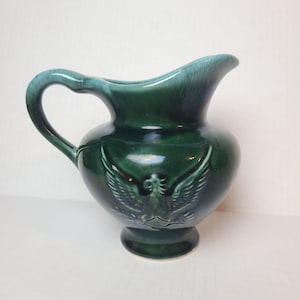May include: A green ceramic pitcher with a raised design of an eagle with outstretched wings. The pitcher has a light green glaze and a slightly iridescent finish.