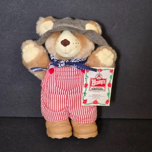 May include: A plush teddy bear wearing a gray hat, a blue bandana, and red and white striped overalls. The bear has tan fur and brown boots. A Wendy's tag is attached to the bear.