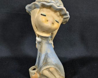 Könnte beinhalten: Keramikfigur eines sitzenden Mädchens in einem blauen Kleid und einem passenden Hut mit Rüschenrand. Die Figur hat geschlossene Augen und einen kleinen Korb. Die Farbpalette umfasst Beige, Blau und einen Hauch von Orange.
