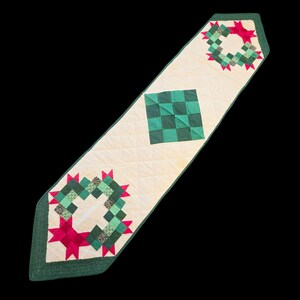 May include: A quilted table runner with an off-white base featuring a central green checkered square and two end motifs composed of small green squares and red star-shaped accents, all bordered by a dark green fabric trim with diamond stitching.