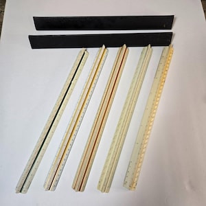 May include: Five vintage rulers with ivory-colored plastic bodies and black markings. The rulers have different colored edges, including green, gold, and black. The rulers are laid out on a white surface.