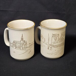 May include: Two beige ceramic mugs with brown line drawings of a church and a rural scene. Each mug has a gold rim and a curved handle. The mugs are likely for coffee or tea.
