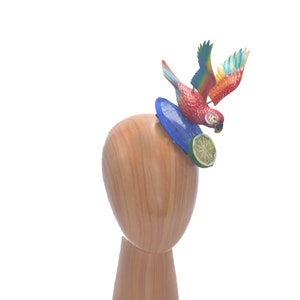 May include: A colorful parrot figurine perched on a blue and white surfboard with a lime wedge. The figurine is mounted on a wooden mannequin head.