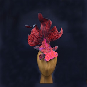 May include: A red and pink goldfish headpiece with black detailing. The headpiece is made of fabric and is designed to look like a goldfish. It is sitting on a wooden mannequin head.