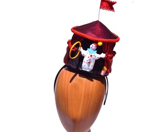 May include: A red and black circus tent hat with a clown figurine inside. The hat is on a wooden mannequin head.