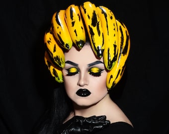 May include: A person with black hair and dark makeup is wearing a crown of yellow bananas with black markings. The person is wearing a black top and black gloves. The background is black. The text "LACEY NOEL 100 DAYS OF MAKEUP 19" is at the bottom of the image.