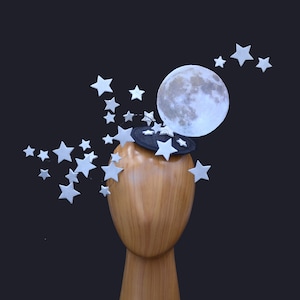 Puede incluir: Una luna de fieltro blanco con una base de fieltro negro y estrellas de fieltro plateado, puesta en una cabeza de maniquí de madera.