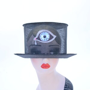 May include: A black mesh top hat with a silver eye clock embellishment. The eye has a blue iris and a pink pupil. The clock face reads "Dalila" and has Roman numerals.