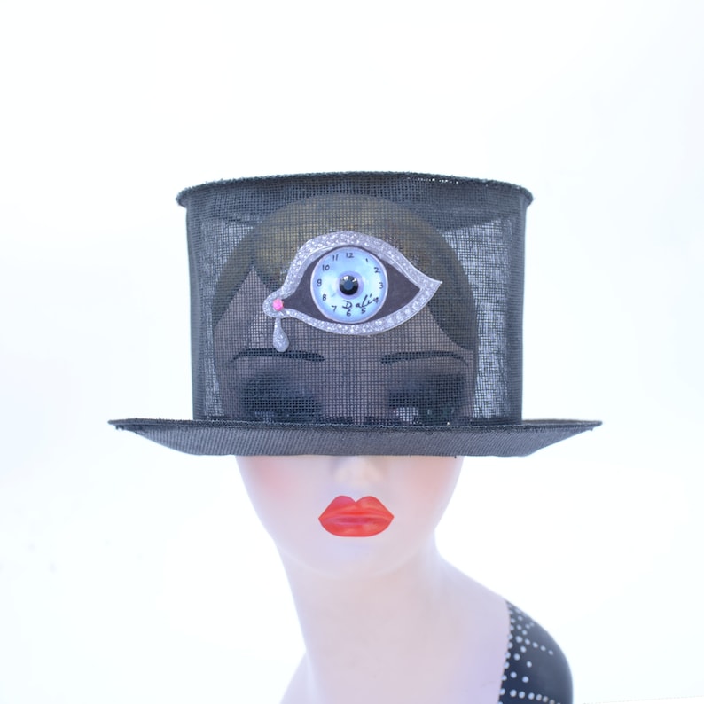 May include: A black mesh top hat with a silver eye clock embellishment. The eye has a blue iris and a pink pupil. The clock face reads "Dalila" and has Roman numerals.