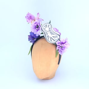 May include: A black headband with a purple and white flower crown and a white paper clock face. The clock face is shaped like a pocket watch and has Roman numerals.