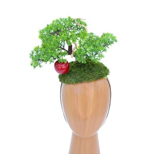 May include: A miniature green tree with a red apple and a gold snake, growing out of a moss-covered base, on a wooden mannequin head.