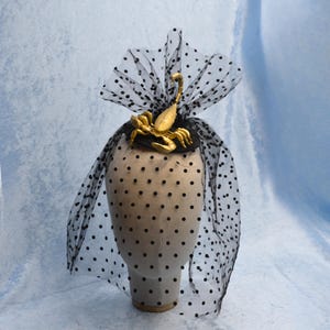 May include: A black fascinator with a gold scorpion embellishment. The hat features a black base and a sheer, dotted veil. The scorpion is the focal point, adding a unique and eye-catching detail to this headpiece.