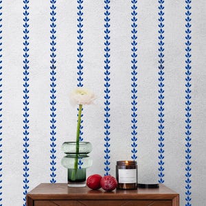 Può includere: Un comodino in legno con un cassetto e un pomello dorato. Sopra, un vaso di vetro verde contiene un fiore bianco. Una candela accesa e due prugne sono accanto al vaso. Il muro dietro è bianco con motivi di foglie blu verticali.