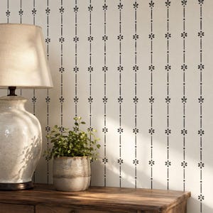 Könnte beinhalten: Eine Wand mit einem sich wiederholenden vertikalen Muster aus schwarzen Linien, kleinen Rautenformen und stilisierten Blumenmotiven auf cremefarbenem Hintergrund. Das Design erzeugt eine strukturierte, lineare Ästhetik.
