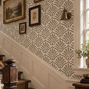 May include: A wall covered in cream-colored wallpaper featuring a repeating dark gray leaf-like fan pattern. The wall is decorated with three framed pictures and a brass wall sconce, positioned above a white wainscoted staircase.